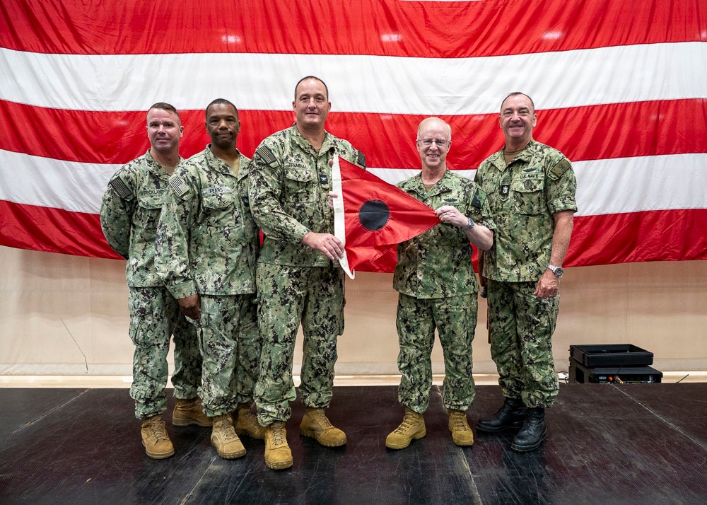Five U.S. Navy personnel in NWU Type III (green camouflage) uniforms pose for a formal group photo on a stage. Two of the central figures are jointly holding a red flag with a black circle, suggesting a change of command or unit ceremony. A large American flag serves as the backdrop.