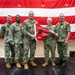 Five U.S. Navy personnel in NWU Type III (green camouflage) uniforms pose for a formal group photo on a stage. Two of the central figures are jointly holding a red flag with a black circle, suggesting a change of command or unit ceremony. A large American flag serves as the backdrop.