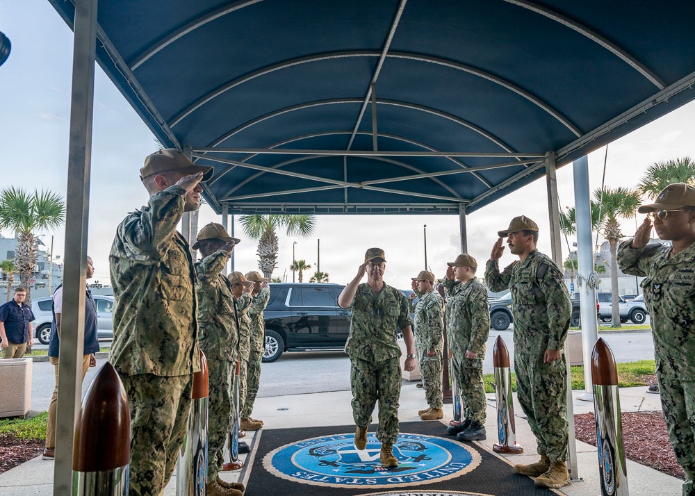 A formal military ceremony is taking place at a U.S. Navy installation, where personnel in Navy Working Uniform (NWU Type III) are rendering salutes to a senior officer walking through a sideboy formation. The scene is set under a covered entrance canopy with a U.S. Navy seal emblem visible on the floor mat. This appears to be an official arrival or departure ceremony, likely for a flag officer or senior dignitary.