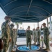A formal military ceremony is taking place at a U.S. Navy installation, where personnel in Navy Working Uniform (NWU Type III) are rendering salutes to a senior officer walking through a sideboy formation. The scene is set under a covered entrance canopy with a U.S. Navy seal emblem visible on the floor mat. This appears to be an official arrival or departure ceremony, likely for a flag officer or senior dignitary.