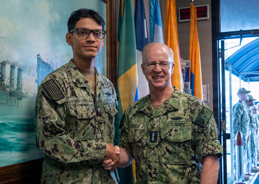 Two U.S. Navy personnel in NWU Type III (AOR-2) camouflage uniforms are shaking hands in what appears to be an official military setting. The individual on the right, identified by name tape as 'CAUDLE,' displays four-star rank insignia indicating the rank of Admiral, while the individual on the left, identified as 'GONZALEZMIRANDA,' appears to be a Petty Officer. The setting includes multiple ceremonial flags and a naval painting in the background, suggesting a formal recognition or award ceremony.