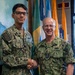 Two U.S. Navy personnel in NWU Type III (AOR-2) camouflage uniforms are shaking hands in what appears to be an official military setting. The individual on the right, identified by name tape as 'CAUDLE,' displays four-star rank insignia indicating the rank of Admiral, while the individual on the left, identified as 'GONZALEZMIRANDA,' appears to be a Petty Officer. The setting includes multiple ceremonial flags and a naval painting in the background, suggesting a formal recognition or award ceremony.