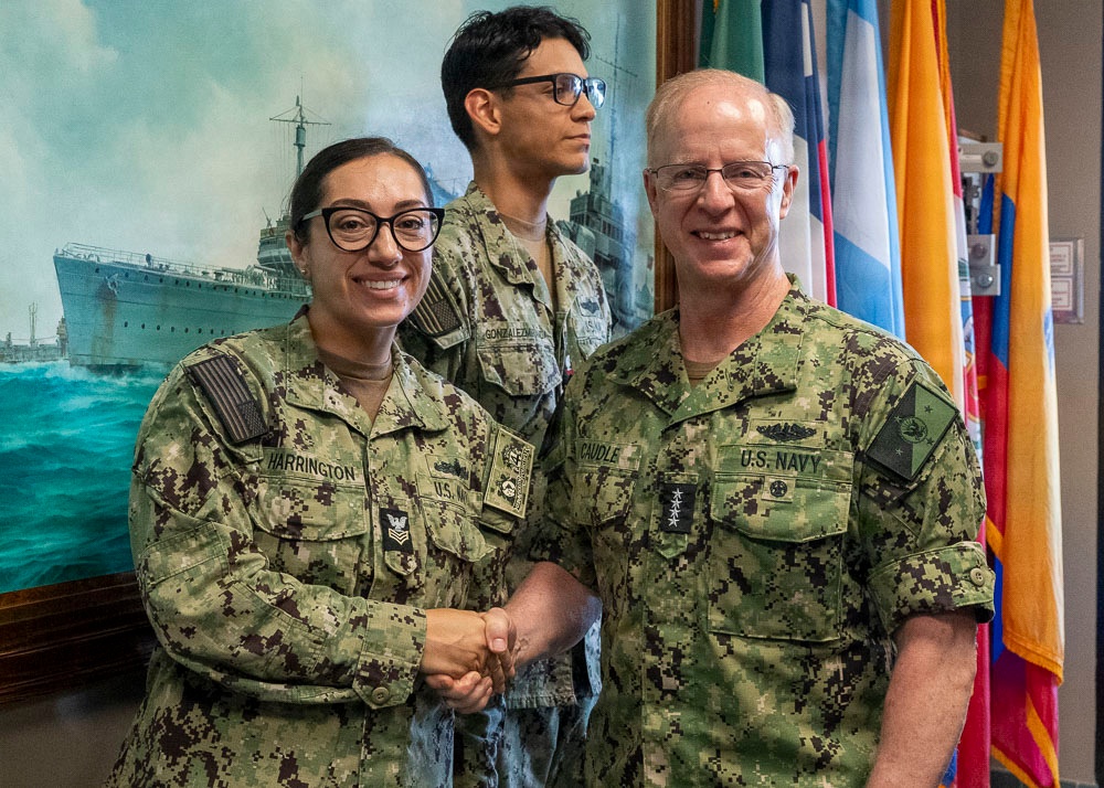 Three U.S. Navy personnel in NWU Type III (digital camouflage) uniforms are gathered in what appears to be a military office or command space. Two individuals in the foreground — name-taped 'Harrington' and 'Caudle' — are shaking hands in what appears to be a formal recognition or ceremony. A naval painting of a ship and multiple flags are visible in the background.