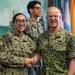 Three U.S. Navy personnel in NWU Type III (digital camouflage) uniforms are gathered in what appears to be a military office or command space. Two individuals in the foreground — name-taped 'Harrington' and 'Caudle' — are shaking hands in what appears to be a formal recognition or ceremony. A naval painting of a ship and multiple flags are visible in the background.
