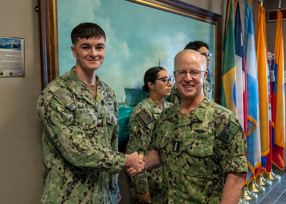 Two U.S. Navy personnel in NWU Type III (digital camouflage) uniforms are shaking hands in what appears to be an official military building or headquarters. The individual on the right displays four stars on their collar, indicating the rank of Admiral, while the individual on the left has the name tape 'WALSH'. Two additional service members are visible in the background.