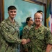 Two U.S. Navy personnel in NWU Type III (digital camouflage) uniforms are shaking hands in what appears to be an official military building or headquarters. The individual on the right displays four stars on their collar, indicating the rank of Admiral, while the individual on the left has the name tape 'WALSH'. Two additional service members are visible in the background.