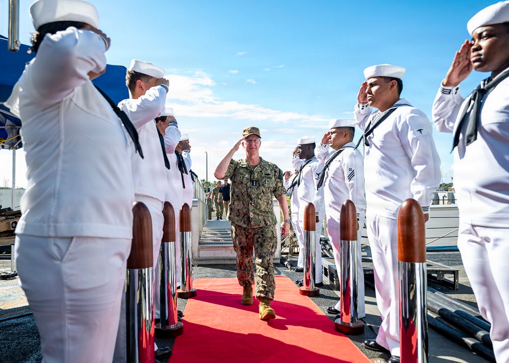 A senior military officer in camouflage uniform walks along a red carpet while being rendered honors by a sideboy formation of U.S. Navy sailors in white dress uniforms. The officer returns the salute as he passes between two rows of sailors flanking chrome-topped stanchions on what appears to be a naval pier or vessel deck. This is a formal military honors ceremony consistent with a high-ranking official's arrival or departure.