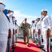 A senior military officer in camouflage uniform walks along a red carpet while being rendered honors by a sideboy formation of U.S. Navy sailors in white dress uniforms. The officer returns the salute as he passes between two rows of sailors flanking chrome-topped stanchions on what appears to be a naval pier or vessel deck. This is a formal military honors ceremony consistent with a high-ranking official's arrival or departure.