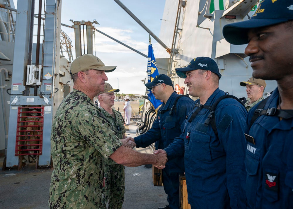 A senior U.S. Navy officer in camouflage utilities and a tan cap is shaking hands with sailors dressed in navy blue coveralls and dark caps, likely during a greeting or departure ceremony aboard a naval vessel. Multiple sailors are lined up in formation, suggesting a formal welcome or farewell event. A blue military unit guidon flag is visible in the background.