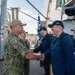 A senior U.S. Navy officer in camouflage utilities and a tan cap is shaking hands with sailors dressed in navy blue coveralls and dark caps, likely during a greeting or departure ceremony aboard a naval vessel. Multiple sailors are lined up in formation, suggesting a formal welcome or farewell event. A blue military unit guidon flag is visible in the background.