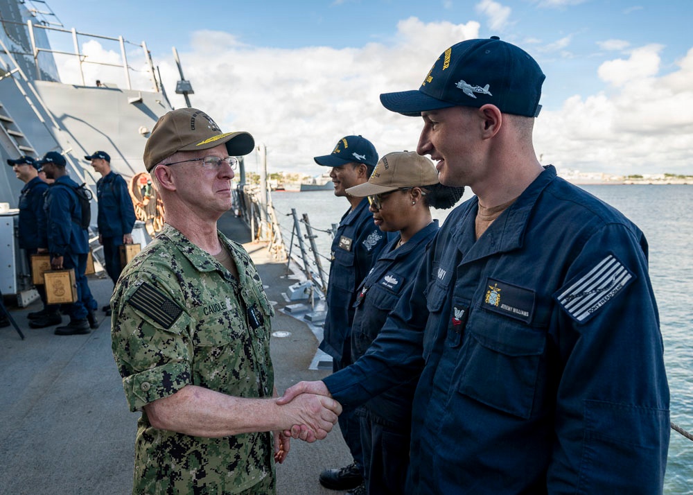 A senior U.S. Navy officer in camouflage utilities and a tan cover is shaking hands with a sailor in navy blue coveralls on the deck of a naval vessel at a pier. The name tape 'CAUDLE' is visible on the officer's uniform, and the sailor being greeted is identified as 'JEREMY WILLIAMS' by his name tape. Several other sailors in blue coveralls and tan covers are lined up in the background, suggesting a formal greeting or inspection event.