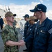 A senior U.S. Navy officer in camouflage utilities and a tan cover is shaking hands with a sailor in navy blue coveralls on the deck of a naval vessel at a pier. The name tape 'CAUDLE' is visible on the officer's uniform, and the sailor being greeted is identified as 'JEREMY WILLIAMS' by his name tape. Several other sailors in blue coveralls and tan covers are lined up in the background, suggesting a formal greeting or inspection event.