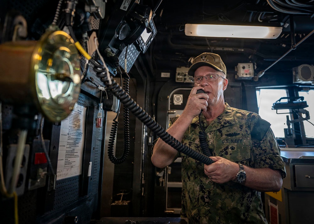 A U.S. Navy officer in digital camouflage uniform is speaking into a handheld radio/intercom handset aboard what appears to be a naval vessel's bridge or command area. The individual is wearing a cover (cap) with what appears to be a star insignia, suggesting flag officer rank. The surrounding environment is filled with communication equipment, mounted panels, and naval instrumentation.