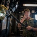 A U.S. Navy officer in digital camouflage uniform is speaking into a handheld radio/intercom handset aboard what appears to be a naval vessel's bridge or command area. The individual is wearing a cover (cap) with what appears to be a star insignia, suggesting flag officer rank. The surrounding environment is filled with communication equipment, mounted panels, and naval instrumentation.