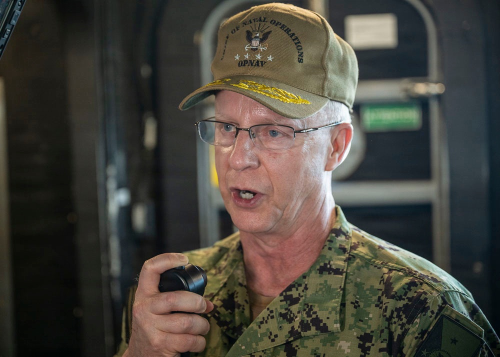 A senior U.S. Navy officer is speaking into a handheld microphone or radio device, wearing a digital camouflage uniform and an olive-colored cap. The cap is embroidered with 'Office of Naval Operations' and 'OPNAV' along with gold leaf insignia, indicating a high-ranking flag officer. The setting appears to be aboard a naval vessel or military facility.