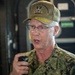 A senior U.S. Navy officer is speaking into a handheld microphone or radio device, wearing a digital camouflage uniform and an olive-colored cap. The cap is embroidered with 'Office of Naval Operations' and 'OPNAV' along with gold leaf insignia, indicating a high-ranking flag officer. The setting appears to be aboard a naval vessel or military facility.