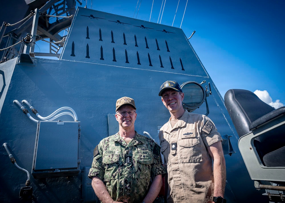 Two military personnel in uniform are posing together on the deck of a naval vessel. The individual on the left wears U.S. Navy digital camouflage (NWU) with a name tape reading 'CAUDLE' and 'U.S. NAVY', while the individual on the right wears a tan/khaki uniform with a name tape reading 'COOK'. Behind them is a large gray superstructure of the ship decorated with numerous kill markings (silhouettes of missiles and aircraft).