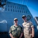 Two military personnel in uniform are posing together on the deck of a naval vessel. The individual on the left wears U.S. Navy digital camouflage (NWU) with a name tape reading 'CAUDLE' and 'U.S. NAVY', while the individual on the right wears a tan/khaki uniform with a name tape reading 'COOK'. Behind them is a large gray superstructure of the ship decorated with numerous kill markings (silhouettes of missiles and aircraft).