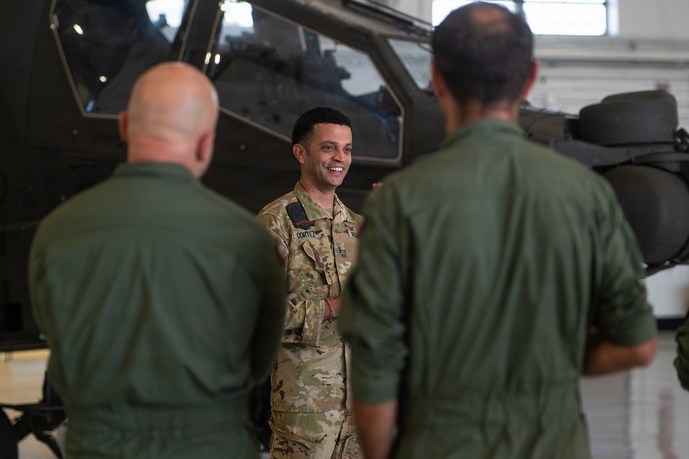 Three military personnel are engaged in conversation inside what appears to be a helicopter hangar. The central figure, wearing U.S. Army OCP (Operational Camouflage Pattern) uniform with a name tape reading 'CORTEZ', is smiling and facing two individuals in olive green flight suits whose backs are to the camera. A military helicopter, consistent with an AH-64 Apache, is visible in the background.