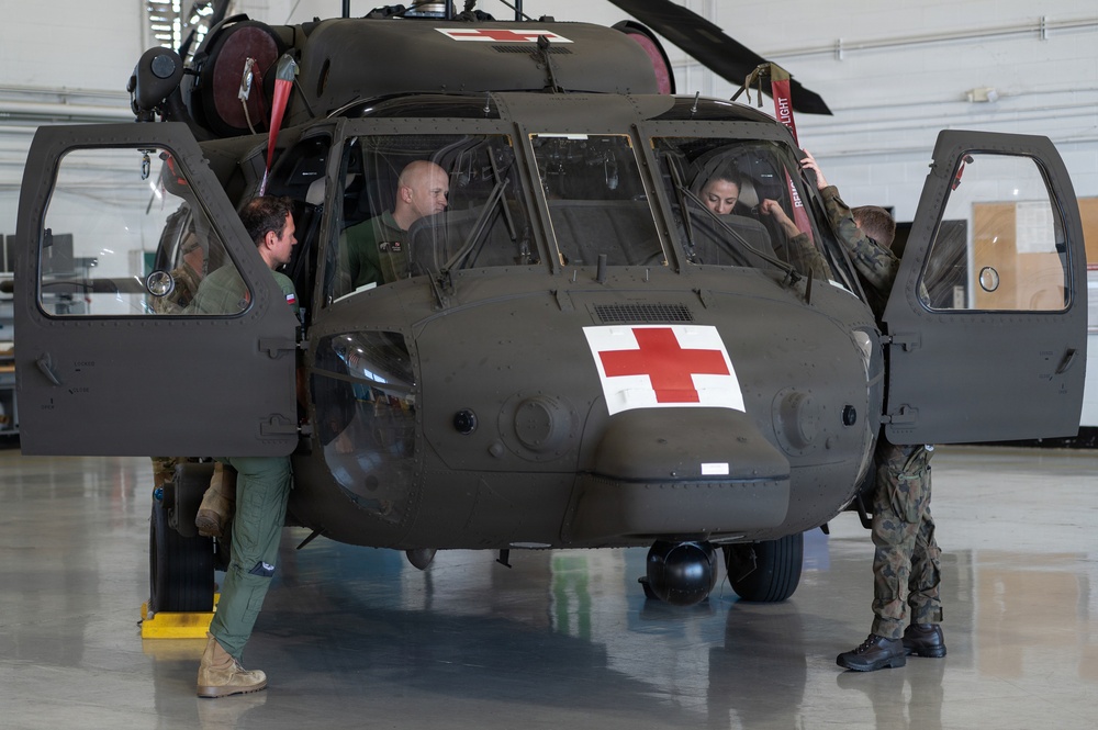 A military medical evacuation (MEDEVAC) helicopter, identifiable by the prominent Red Cross markings, is being inspected or demonstrated inside a hangar facility. Multiple personnel in military uniforms are gathered around and inside the olive-drab helicopter, suggesting a training, familiarization, or pre-flight inspection activity. The aircraft appears to be a UH-60 Black Hawk configured for medical evacuation missions.