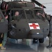 A military medical evacuation (MEDEVAC) helicopter, identifiable by the prominent Red Cross markings, is being inspected or demonstrated inside a hangar facility. Multiple personnel in military uniforms are gathered around and inside the olive-drab helicopter, suggesting a training, familiarization, or pre-flight inspection activity. The aircraft appears to be a UH-60 Black Hawk configured for medical evacuation missions.
