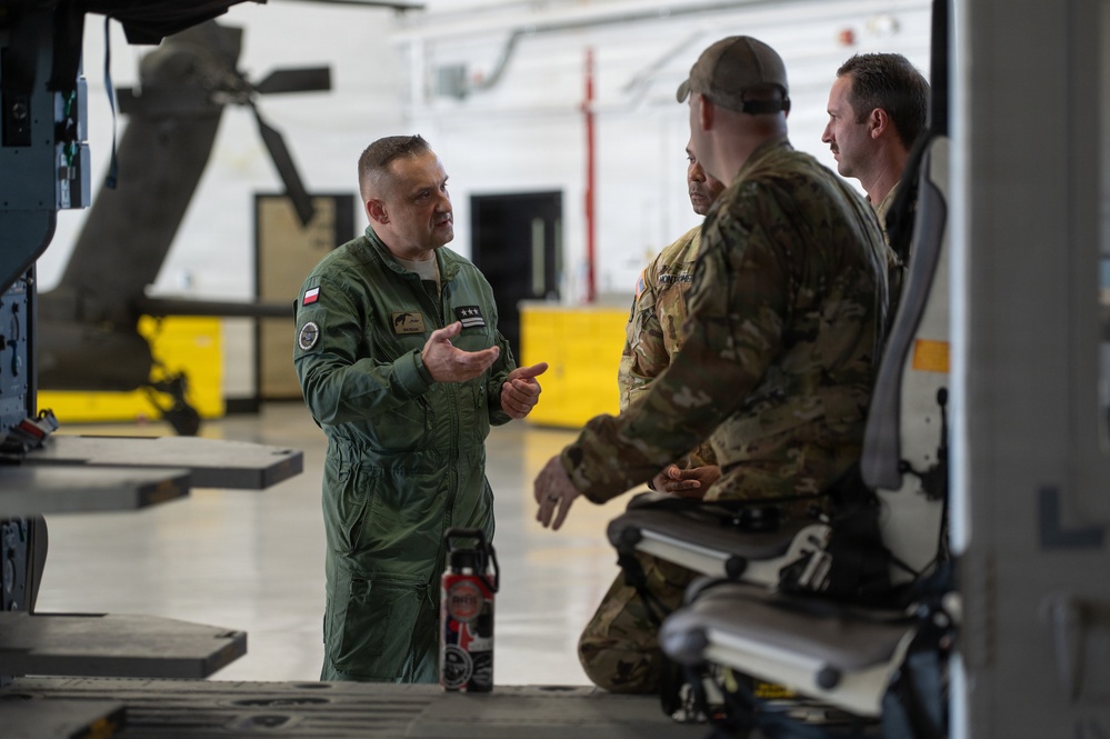 Three military personnel are engaged in conversation inside what appears to be an aircraft hangar. The individual on the left is wearing a green flight suit with a Polish flag patch and two-star rank insignia, speaking to two others in camouflage uniforms. A helicopter or large aircraft is partially visible in the background.