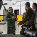 Three military personnel are engaged in conversation inside what appears to be an aircraft hangar. The individual on the left is wearing a green flight suit with a Polish flag patch and two-star rank insignia, speaking to two others in camouflage uniforms. A helicopter or large aircraft is partially visible in the background.