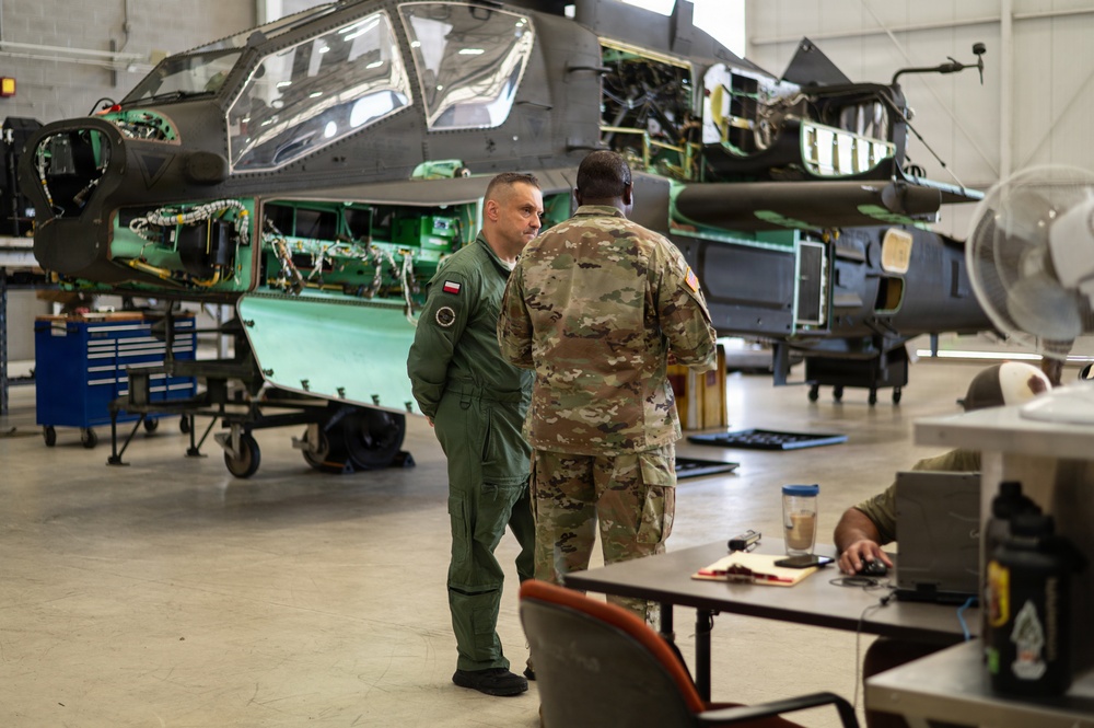 Two military personnel are conversing inside a maintenance hangar in front of a partially disassembled military attack helicopter, likely an AH-64 Apache. One individual wears an olive green flight suit with what appears to be a Polish flag patch, while the other wears US Army OCP (Operational Camouflage Pattern) uniform. A third person is seated at a desk working on a computer in the foreground.