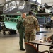Two military personnel are conversing inside a maintenance hangar in front of a partially disassembled military attack helicopter, likely an AH-64 Apache. One individual wears an olive green flight suit with what appears to be a Polish flag patch, while the other wears US Army OCP (Operational Camouflage Pattern) uniform. A third person is seated at a desk working on a computer in the foreground.