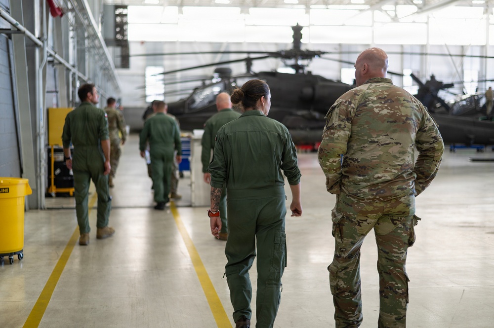 Military personnel are walking through a large aircraft hangar containing what appear to be Apache attack helicopters. The group consists of individuals wearing green flight suits and one person in MultiCam/OCP camouflage uniform. The setting appears to be an active military aviation maintenance or inspection facility.