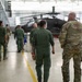 Military personnel are walking through a large aircraft hangar containing what appear to be Apache attack helicopters. The group consists of individuals wearing green flight suits and one person in MultiCam/OCP camouflage uniform. The setting appears to be an active military aviation maintenance or inspection facility.