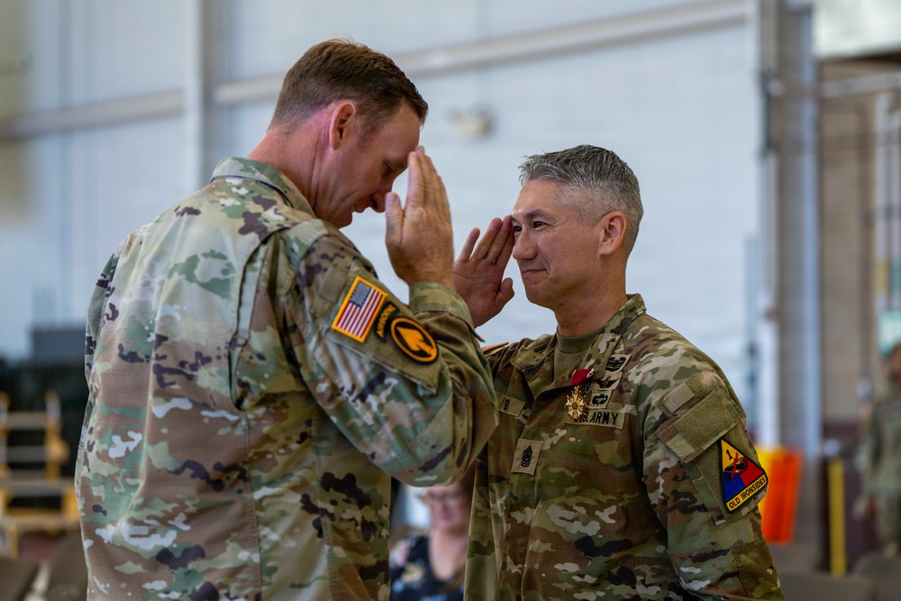 Two U.S. Army soldiers in OCP (Operational Camouflage Pattern) uniforms are exchanging salutes in what appears to be a military ceremony inside a hangar or large facility. The soldier on the right is wearing a medal on his chest, suggesting this is a decoration or awards ceremony. Both soldiers are facing each other in a formal military salute.
