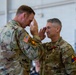 Two U.S. Army soldiers in OCP (Operational Camouflage Pattern) uniforms are exchanging salutes in what appears to be a military ceremony inside a hangar or large facility. The soldier on the right is wearing a medal on his chest, suggesting this is a decoration or awards ceremony. Both soldiers are facing each other in a formal military salute.
