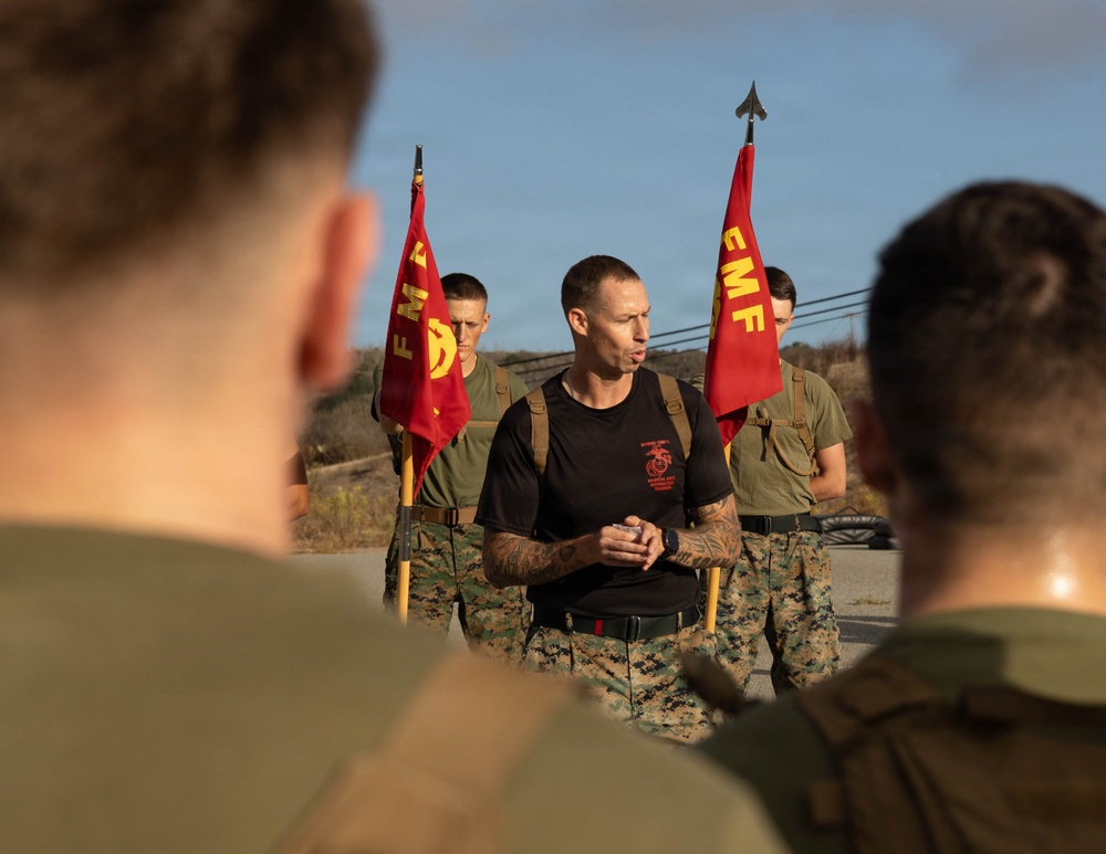 A U.S. Marine Corps leader, wearing a black t-shirt with Marine Corps insignia and camouflage trousers, addresses a group of Marines in an outdoor setting. Two red FMF (Fleet Marine Force) guidon flags with the Marine Corps Eagle, Globe, and Anchor emblem are held by flag bearers flanking the central figure. The scene appears to be a pre-event briefing or formation address.