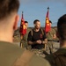 A U.S. Marine Corps leader, wearing a black t-shirt with Marine Corps insignia and camouflage trousers, addresses a group of Marines in an outdoor setting. Two red FMF (Fleet Marine Force) guidon flags with the Marine Corps Eagle, Globe, and Anchor emblem are held by flag bearers flanking the central figure. The scene appears to be a pre-event briefing or formation address.