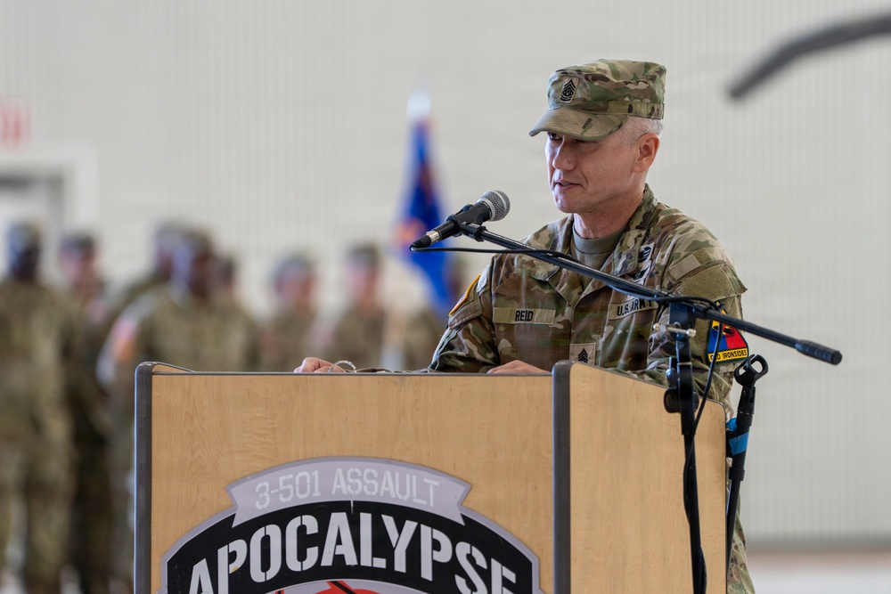 A U.S. Army soldier, identified by name tape as 'REID', stands at a wooden podium speaking into a microphone at what appears to be a military ceremony. The podium bears the insignia and text '3-501 ASSAULT APOCALYPSE'. The soldier wears the Operational Camouflage Pattern (OCP) uniform with sergeant major rank insignia and what appears to be an 'Old Ironsides' (1st Armored Division) patch.