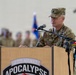 A U.S. Army soldier, identified by name tape as 'REID', stands at a wooden podium speaking into a microphone at what appears to be a military ceremony. The podium bears the insignia and text '3-501 ASSAULT APOCALYPSE'. The soldier wears the Operational Camouflage Pattern (OCP) uniform with sergeant major rank insignia and what appears to be an 'Old Ironsides' (1st Armored Division) patch.