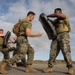 A group of military personnel, appearing to be U.S. Marines based on their MARPAT camouflage uniforms, are engaged in a close-combat or martial arts training exercise on a paved outdoor surface. Trainees are using large black padded shields during what appears to be a striking or combatives drill. The image is taken from a low angle, capturing dynamic movement with some motion blur on the foreground figure.
