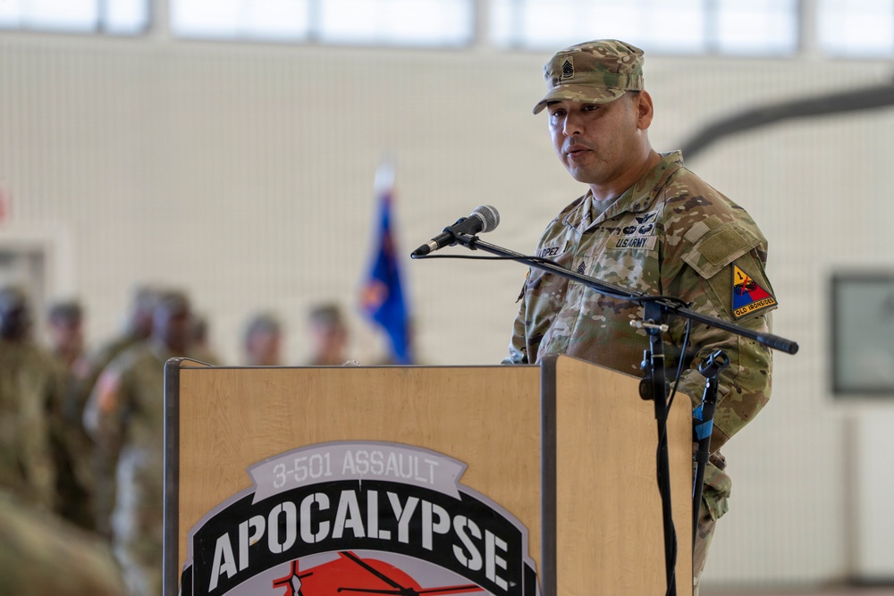 A U.S. Army soldier, identifiable as a senior NCO, stands at a wooden podium bearing the '3-501 Assault Apocalypse' unit insignia and is addressing an audience in what appears to be a military hangar. The soldier is wearing the Operational Camouflage Pattern (OCP) uniform with visible rank insignia, a name tape reading 'LOPEZ,' and a shoulder patch consistent with the 1st Armored Division ('Old Ironsides'). A formation of soldiers and a unit flag are visible in the blurred background.