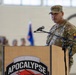 A U.S. Army soldier, identifiable as a senior NCO, stands at a wooden podium bearing the '3-501 Assault Apocalypse' unit insignia and is addressing an audience in what appears to be a military hangar. The soldier is wearing the Operational Camouflage Pattern (OCP) uniform with visible rank insignia, a name tape reading 'LOPEZ,' and a shoulder patch consistent with the 1st Armored Division ('Old Ironsides'). A formation of soldiers and a unit flag are visible in the blurred background.