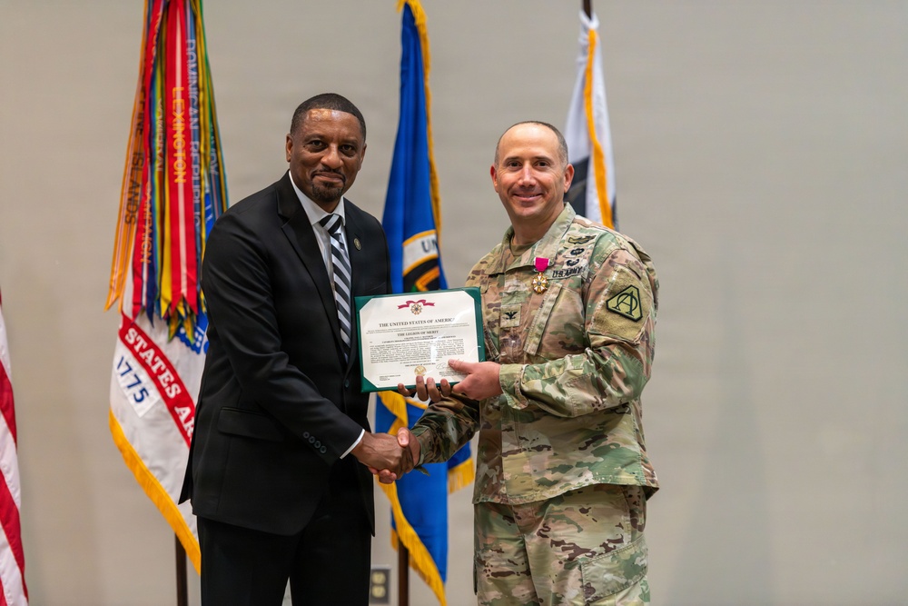 Two individuals are posing together during a formal military award ceremony. A civilian in a dark suit and a U.S. Army soldier in OCP (Operational Camouflage Pattern) uniform are jointly holding a Legion of Merit certificate while shaking hands. The soldier is wearing the Legion of Merit medal on his chest, indicating he is the award recipient.