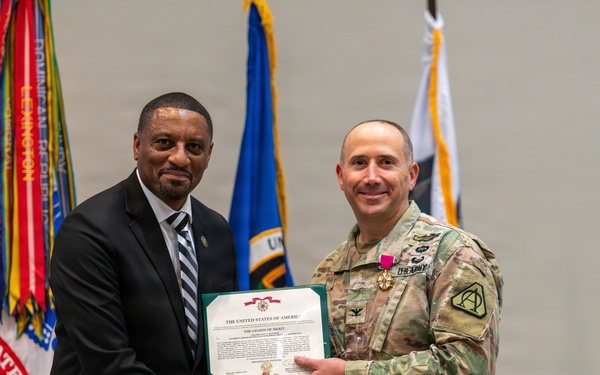 Two individuals are posing together during a formal military award ceremony. A civilian in a dark suit and a U.S. Army soldier in OCP (Operational Camouflage Pattern) uniform are jointly holding a Legion of Merit certificate while shaking hands. The soldier is wearing the Legion of Merit medal on his chest, indicating he is the award recipient.
