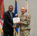 Two individuals are posing together during a formal military award ceremony. A civilian in a dark suit and a U.S. Army soldier in OCP (Operational Camouflage Pattern) uniform are jointly holding a Legion of Merit certificate while shaking hands. The soldier is wearing the Legion of Merit medal on his chest, indicating he is the award recipient.