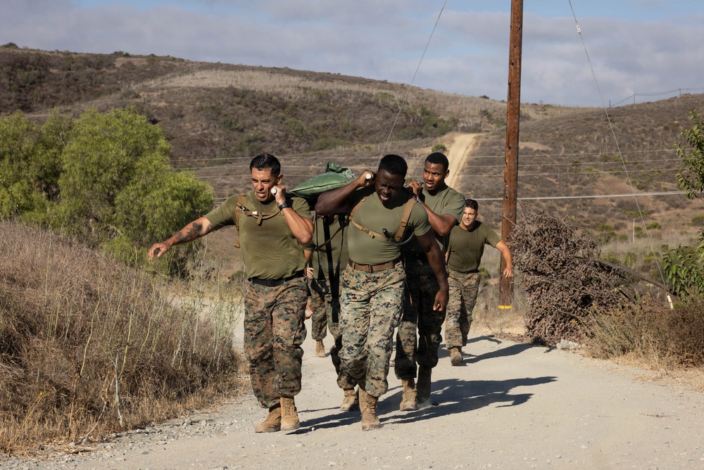 A group of military personnel in U.S. Marine Corps uniforms are conducting a physical training exercise on a dirt road in a dry, hilly terrain. Two lead individuals are carrying a heavy green sandbag or loaded pack across their shoulders while others follow closely behind. The setting appears to be a training base, likely in Southern California based on the landscape.