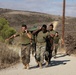 A group of military personnel in U.S. Marine Corps uniforms are conducting a physical training exercise on a dirt road in a dry, hilly terrain. Two lead individuals are carrying a heavy green sandbag or loaded pack across their shoulders while others follow closely behind. The setting appears to be a training base, likely in Southern California based on the landscape.