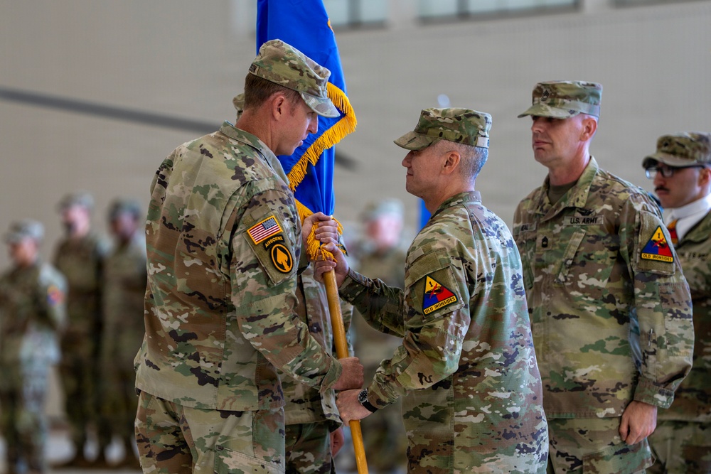 A military change of command ceremony is taking place indoors, likely in a hangar or gymnasium. Two senior officers are exchanging a unit guidon (flag), which is the central symbolic act of a change of command ceremony. Additional soldiers in U.S. Army OCP (Operational Camouflage Pattern) uniforms are visible in the background.