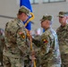 A military change of command ceremony is taking place indoors, likely in a hangar or gymnasium. Two senior officers are exchanging a unit guidon (flag), which is the central symbolic act of a change of command ceremony. Additional soldiers in U.S. Army OCP (Operational Camouflage Pattern) uniforms are visible in the background.