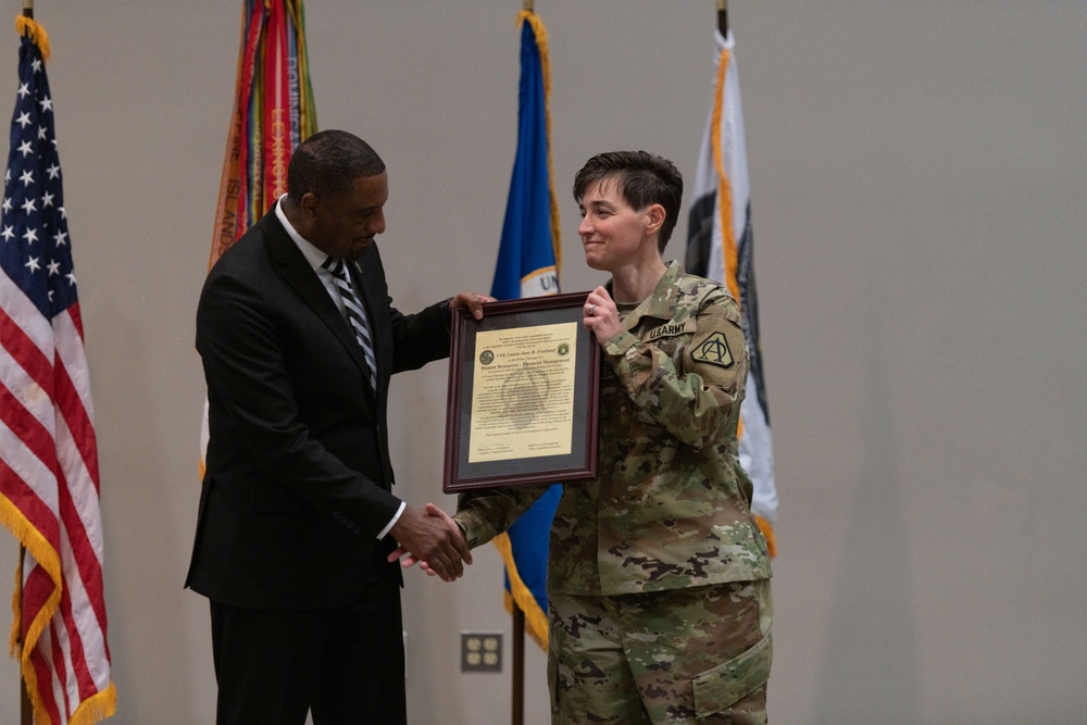 A formal military ceremony in which a civilian official in a dark suit presents a framed certificate or award to a U.S. Army soldier in OCP (Operational Camouflage Pattern) uniform. The two individuals are shaking hands while jointly holding the framed document. Several flags, including the U.S. flag and military unit flags, are displayed in the background.
