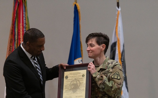 A formal military ceremony in which a civilian official in a dark suit presents a framed certificate or award to a U.S. Army soldier in OCP (Operational Camouflage Pattern) uniform. The two individuals are shaking hands while jointly holding the framed document. Several flags, including the U.S. flag and military unit flags, are displayed in the background.