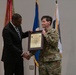 A formal military ceremony in which a civilian official in a dark suit presents a framed certificate or award to a U.S. Army soldier in OCP (Operational Camouflage Pattern) uniform. The two individuals are shaking hands while jointly holding the framed document. Several flags, including the U.S. flag and military unit flags, are displayed in the background.